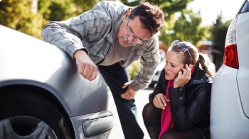 A couple check the damage to the front of their silver car while the woman calls her State Farm agent.